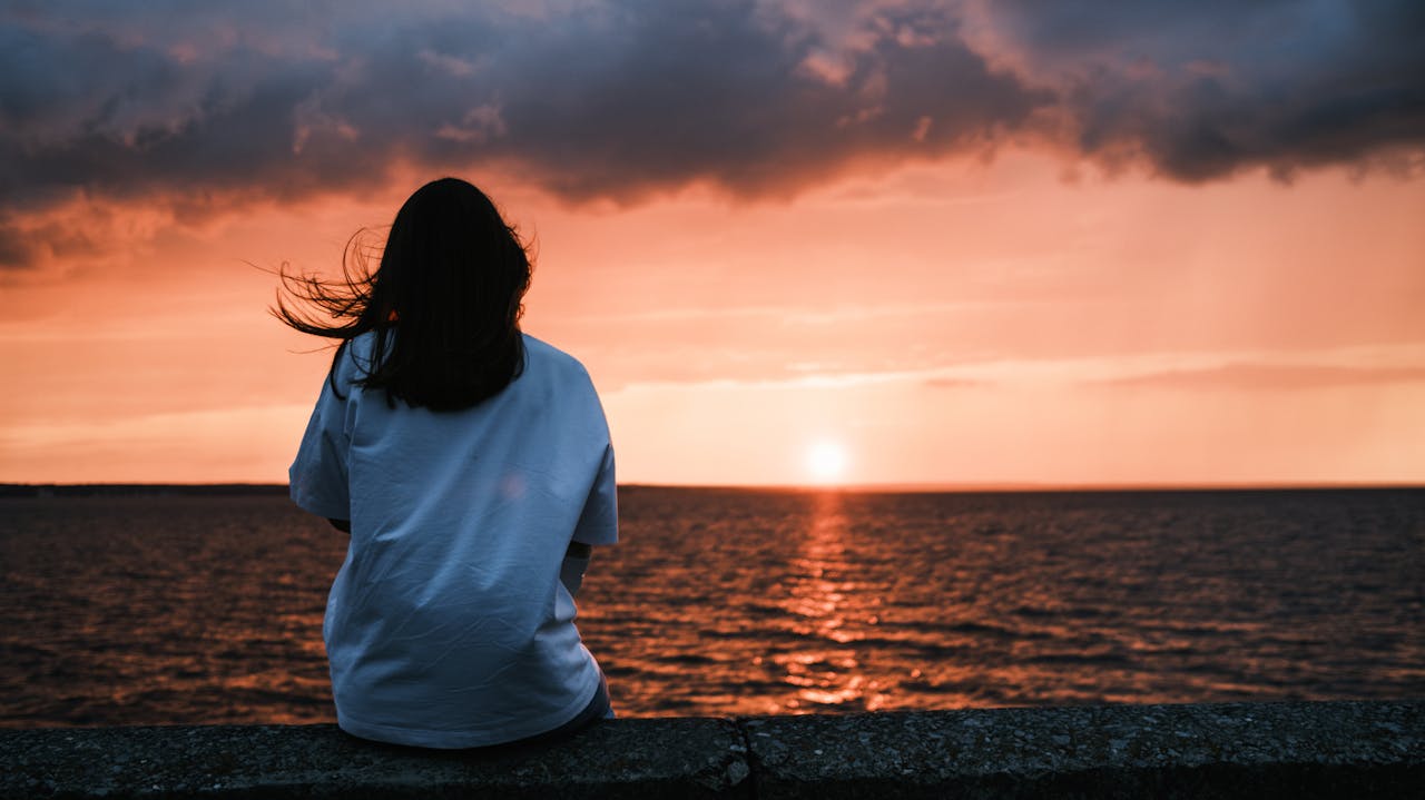 Tranquil evening scene with a woman gazing at a stunning sunset over the ocean, capturing a moment of serenity.